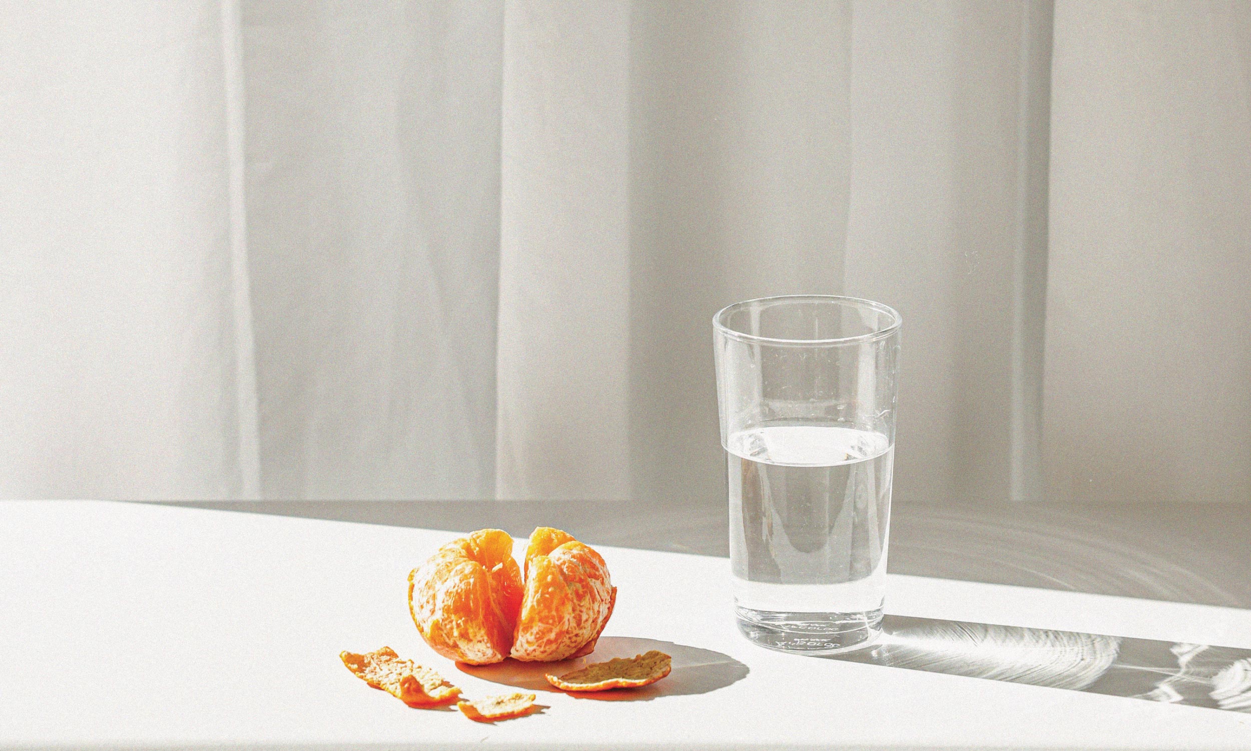 Glass of water beside a peeled orange in sunlight, illustrating the hydration and energy benefits of healthy daily habits