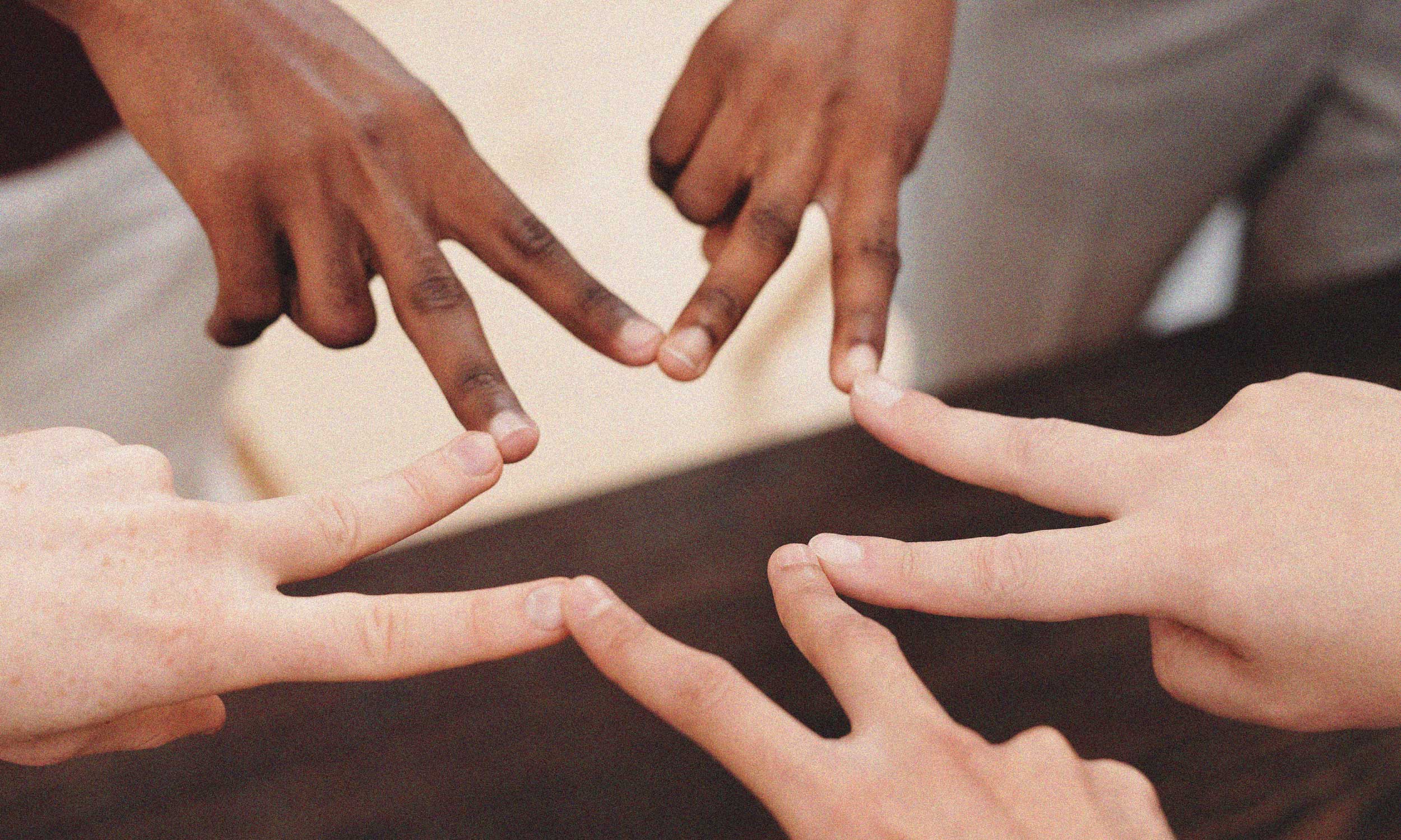 five people of different skin tones extend their index fingers toward the center of the frame, with each fingertip touching another to form a star. The close-up composition focuses on their hands against a softly blurred indoor background, symbolizing unity, mutual respect, equality, and human connection.
